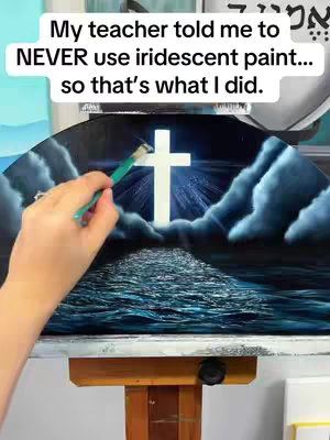 A hand with a paintbrush is applying paint to a canvas depicting a cross emitting light, positioned above a body of water under stylized clouds.