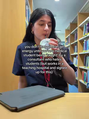 A young woman sits at a desk in a library or similar setting, holding a beverage and looking toward the camera. She appears to be in a medical school setting, with books visible on shelves behind her. Her expression suggests a mixture of weariness and contemplation.