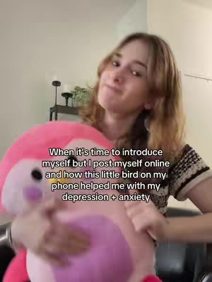 A young woman is holding a large pink plush bird toy in front of her. The woman is smiling and looking towards the camera. Text overlayed on the image reads: "When it's time to introduce myself but I post myself online and how this little bird on my phone helped me with my depression + anxiety". The background is a light-colored wall with some home decor visible.