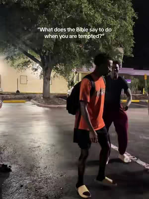 Two men in a parking lot at night. One is wearing an orange shirt and black shorts with a backpack slung over one shoulder. The other is wearing a black shirt and maroon pants. The setting has a tree and buildings in the background with street lights.