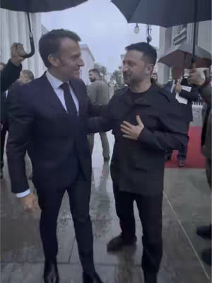 Two men in suits and coats are standing outdoors under umbrellas, two other people are also under umbrellas. One of them has his hand on the other person's arm.