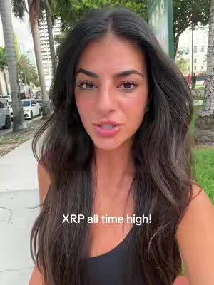 A woman with long dark hair stands outside, looking directly at the camera. She's wearing a dark tank top. Behind her, a walkway, trees, and some buildings are visible, creating a natural outdoor setting.