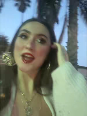 A woman with dark hair and red lipstick looks directly at the camera with an open mouth. She's wearing large gold earrings and a white sweater-like garment over a pink top. The background is blurred, suggesting an outdoor setting.