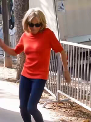 A woman in an orange top and dark pants dances outdoors. She wears sunglasses and moves with a lighthearted expression. A metal fence and a building are in the background.