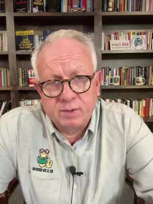 A man in glasses and a light blue shirt is centered in the frame, with a backdrop of bookshelves filled with books. There is a microphone clipped to his shirt.
