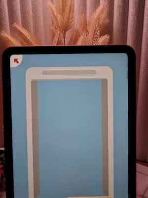 An iPad screen is the focus, displaying a game interface with concentric rectangular frames of white within a blue background. The top of the image includes decoratative elements slightly out of focus.