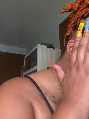 The image is a selfie of a person, likely a woman, who appears to be covering part of their face with their hand. The individual has orange-tinted locs with some pieces gathered at the top. The nails are long and colorful with designs in particular, and nail polish on some of the nails. The upper part of the person’s body is visible, with a black top and a delicate necklace. The background is dimly lit, with a view of a couple of cabinet or boxes and the surrounding room. The overall composition is focused on the person's face and upper body, with a slightly out-of-focus background.