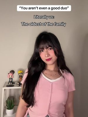 A woman with dark hair and bangs is centered in the image. She wears a light pink button-down shirt, and her lips are a deep red. Behind her, to her left, is a white shelf with small figurines; a green plant in a pot sits below. Text overlays frame the top of the image in white text against a light background.