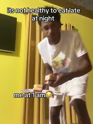 A man in a white t-shirt and shorts is holding a plate of sandwiches in front of a yellow wall. Text overlays read 'its not healthy to eat late at night' and 'me at 1 am'.