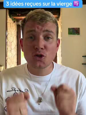A male content creator is centered in the shot, with text overlaid at the top. He is wearing a white t-shirt and a silver necklace. The background includes a wooden-framed window and a painting on the wall.