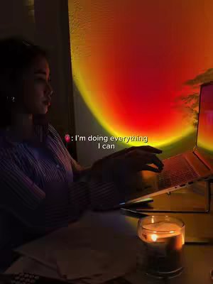 A woman is working on a laptop at a desk, with a candle and papers near the bottom. A sunset lamp provides a warm light behind her.