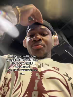 A selfie-style shot featuring a young Black man looking up towards the camera with a slightly pensive expression. He's wearing a black beanie, pink headphones, and a white shirt with a bold graphic print. The hand of another person is seen partially above the frame, seemingly touching the man's head. Text, likely a quote or a caption, is overlaid on the image.