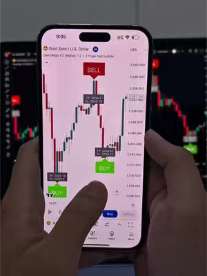 A person's hands holding a smartphone displaying a trading app interface. Overlaid on the phone's screen appears a line graph of data, with 'SELL' and 'BUY' indicators. Dark monitors are visible in the background.
