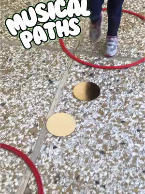 The cover image shows a child's legs and feet wearing sneakers, walking on a floor with beige, speckled tiles. There are multiple red hoops and beige cardboard circles on the floor. Large white text with a bubble effect overlay at the top saying "MUSICAL PATHS".