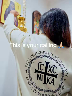 The image shows a person from the back, wearing a cream-colored sweatshirt. The back design features a black cross emblem encircled by a thorny wreath, with text in a circular pattern around it and the words "LORD, LET YOUR WILL BE DONE" at the bottom. The person is holding a lit object (possibly a candle) near a golden object on the left. The background includes a blurred interior with religious imagery. The hook overlaid on the image reads "This is your calling" with prayer hands emoji.