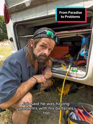 A man kneels in front of a van with the back door open. He is wearing a hat and sunglasses, and he is looking at the camera with a concerned expression. The van's interior is visible, filled with equipment and wires. Text overlay indicates that he is having battery problems.