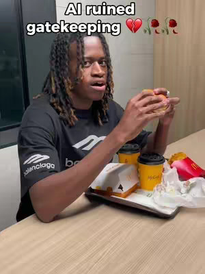 A young Black man with long dreadlocks sits at a table in a fast-food restaurant. He’s holding a hamburger and looking surprised. On the table are McDonald's food items, including fries, coffee cups, and a box with a burger. The background is plain, with a simple wall in the back.