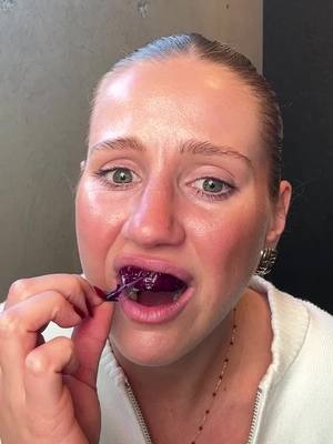A close-up shot of a woman holding a dark purple strip in her mouth, as if she is testing or trying a new product. The background is a neutral gray, and the focus is on her face, with a white top and a delicate gold necklace. Her expression is exaggerated with wide eyes and a slightly open mouth, showing a clear reaction.