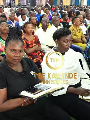 Two women are seated in the foreground of a gathering, possibly a church service or public event. Both are facing forward, with the woman on the left holding a book and a phone. Behind them, a crowd of people of various age and ethnicity are visible. A golden emblem with text is placed in the center of the image.