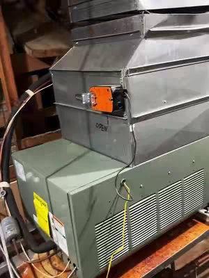 A close-up shot of HVAC equipment, specifically a condenser unit. The unit is composed of multiple sections, with a large rectangular base in a green-gray color and a stacked, metal-paneled structure on top. An orange control element is visible with the word "OPEN" on a metal panel. Wiring and pipes extend from the unit, suggesting an installation or maintenance context. The background suggests an attic environment, with exposed beams and dark shadows.