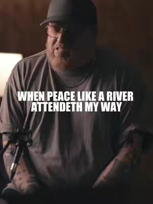 The cover image features a man with a beard wearing a cap and glasses, and a short text overlay. The man is captured from the chest up, with a microphone visible. The lighting appears to be dim, casting shadows, and the background is blurred, suggesting an indoor setting. The man has a neutral facial expression. The color palette is muted with dark tones.