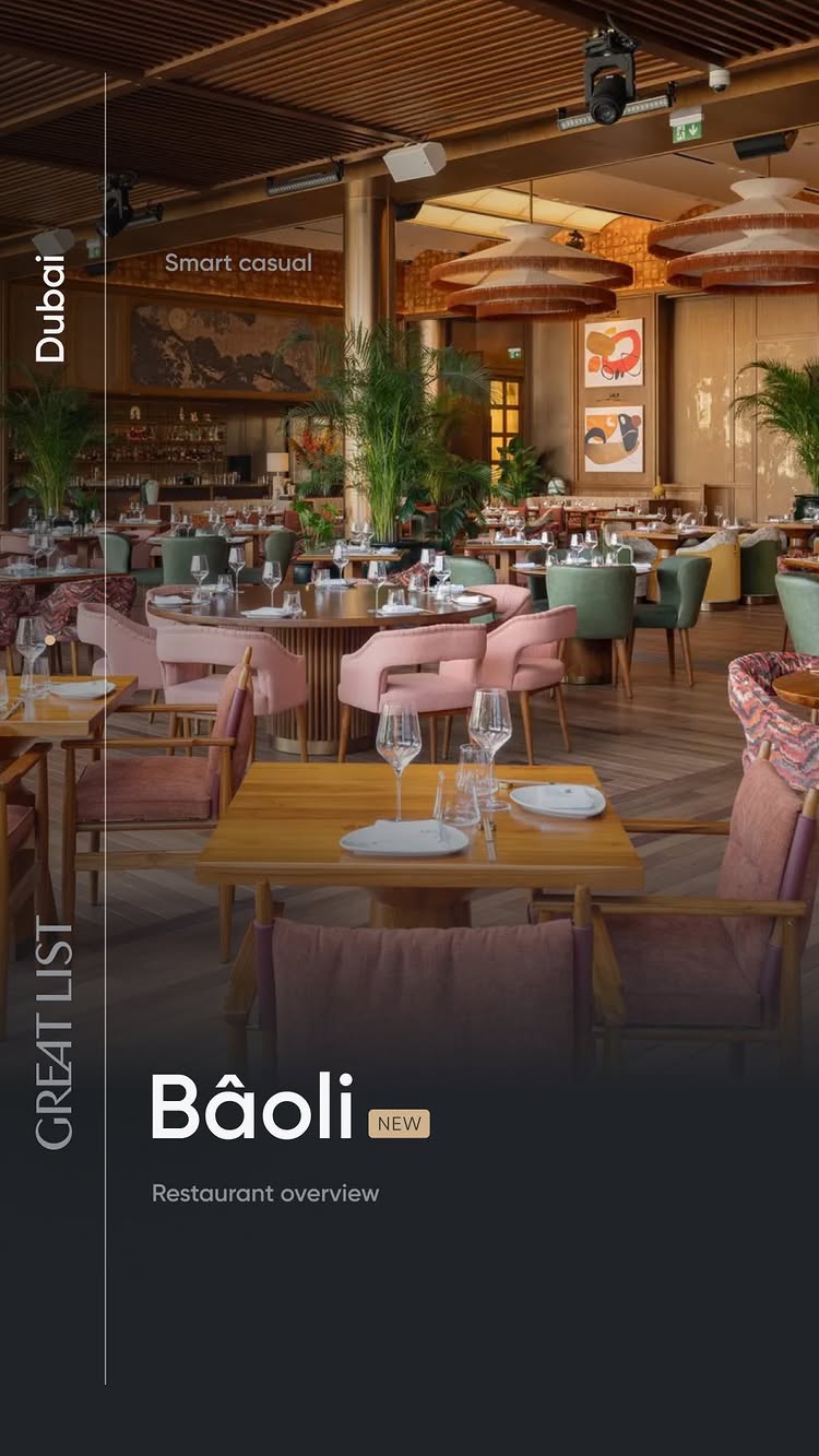 The image showcases the interior of the Bâoli Dubai restaurant. The setting is modern and elegant, with tables set for dining, soft pink and green chairs, and decorative ceiling fixtures. The color palette is warm, with shades of wood, pink, green, and gold.