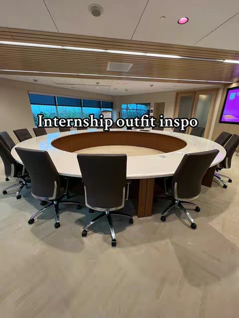 A well-lit, modern conference room setting featuring a large, oval table surrounded by black office chairs. The table has a bright white surface with a central sunken area. Behind, large windows offer a view of the outside. The scene is clean and professional.