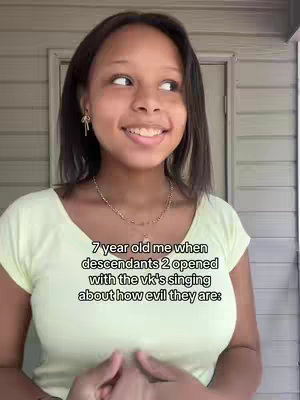 A young woman is smiling while looking slightly upward and to her right. She is wearing a light yellow shirt and a gold necklace. Text overlaid on the image reads: 7 year old me when descendants 2 opened with the vk's singing about how evil they are: