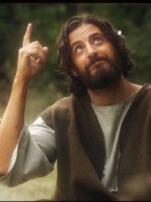 A man with long brown hair and beard, dressed in simple garments, points upwards with his right index finger, with a blurred green backdrop.