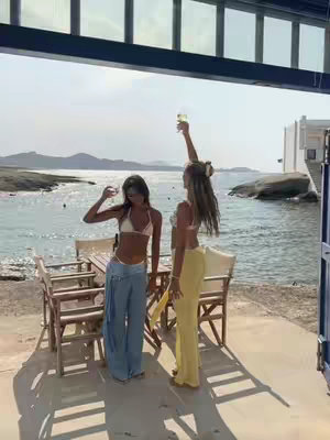 Two young women stand on a concrete patio overlooking a tranquil body of water and distant mountains under a bright sky. Both are wearing light-colored tops and trousers, with one raising a glass in celebration. The scene is framed partially by a dark, architectural structure at the top. The overall composition gives a sense of leisure and warmth.