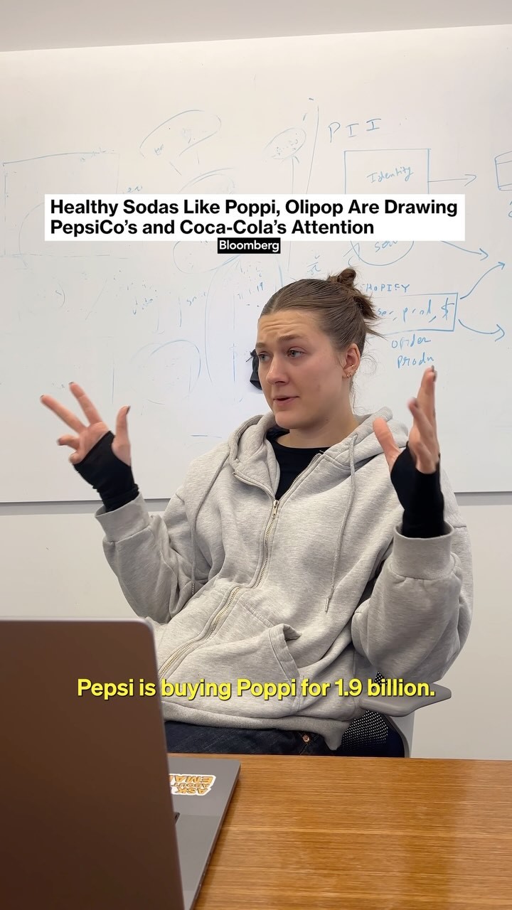 A woman is seated at a desk in front of a whiteboard. The scene has a casual, informational tone, with text overlays providing context about a business deal involving healthy sodas. There is a laptop in front of her. She is gesturing with both hands, looking directly at the camera.