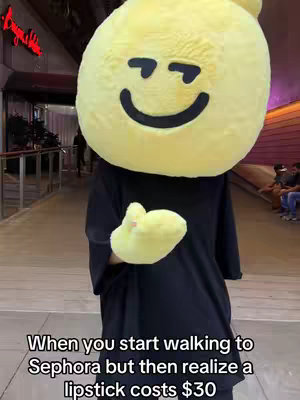 A person wearing a large, yellow, smiling face costume stands on a walkway. The individual is wearing a black outfit. The background shows a blurred out walkway with other people and a store front.