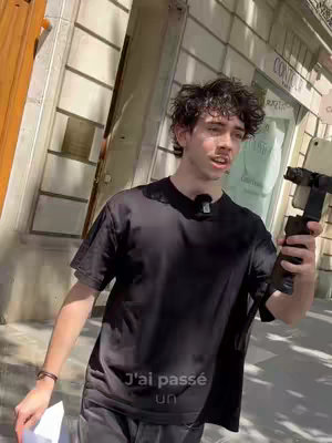 A young man with curly hair stands outdoors, holding a smartphone on a stabilizer. He wears a black t-shirt, and the setting appears to be a city street with stone buildings in the background. Text is overlaid at the bottom of the image.