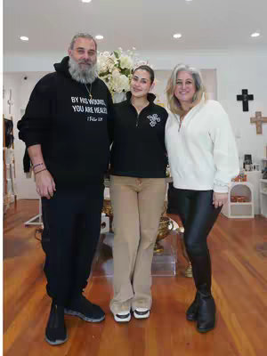 Three people standing in a shop. The man on the left is wearing a black hoodie with text, the woman in the middle is wearing a black top and tan pants, and the woman on the right is wearing a white top and leggings.