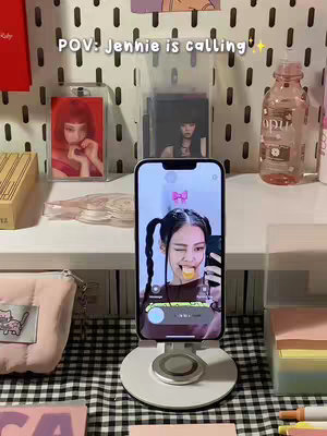 The image shows a phone displaying a selfie of Jennie from Blackpink, on a white stand. The phone's screen shows Jennie with pigtails winking and sticking her tongue out. Behind the phone, there are multiple items on a desk, including pictures, stationary items and a bottle.
