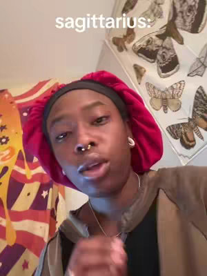 A person with dark skin is centered in the frame. The person is wearing a black beanie, a red cap, and a dark or black top. They have a nose ring. The background includes a colorful patterned cloth and what appears to be a print with butterflies.