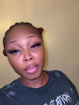A close-up shot of a Black woman against a yellow background. She has braided hair with two small buns on top. Her eyes are closed, with long eyelashes with visible eye makeup. She's wearing a black t-shirt with a graphic, and pale pink lip gloss