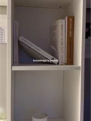 A photo of a white shelf with several books. The shelf is divided into two sections. In the top section, a collection of books stands on their spines, with one book leaning against the others. Below the books, centered, are some white objects, possibly cups or small containers. The lighting appears to be soft and coming from the sides. The text "knowledge is power" is overlaid on the image, and the overall tone is calm and focused.
