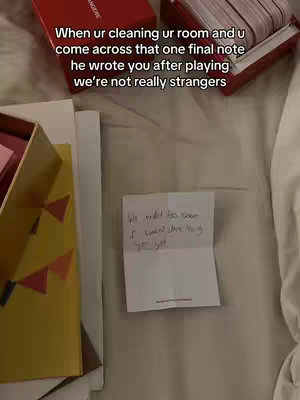The image shows a handwritten note lying on a white surface, likely a bed. Above this is a red box open with message cards. The text overlay says, "When ur cleaning ur room and u come across that one final note he wrote you after playing we're not really strangers."
