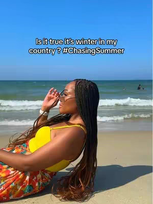 A woman leans on a beach, wearing sunglasses, a yellow top and floral skirt, with the ocean and sky in the background. Text overlay reads: 'Is it true it's winter in my country? #ChasingSummer'
