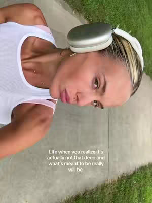 A selfie-style shot. The creator is on the right side of the frame with the head tilted towards the left side, which gives space for the caption. They are outdoors on a sidewalk near green grass. The creator wears a white tank top and silver headphones. The background includes a residential street and sidewalk.
