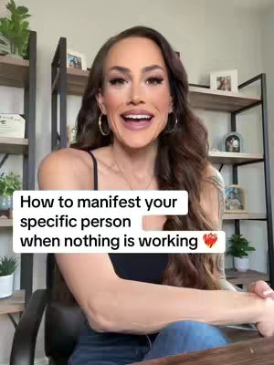 A woman with long brown hair is centered in the frame, smiling and looking at the camera. She's wearing a black top. Behind her, there's a blurred background with shelves and decorative items, suggesting an indoor setting. Text overlay appears on top of the image.
