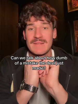 The image shows a man in a dark setting, likely indoors, framed from the chest up. He has dark, curly hair and a large mustache. He wears a black jacket over a white shirt and a silver wristwatch. His hands are clasped in front of him, and he is looking slightly upwards towards the camera. Below the man, there is white text.