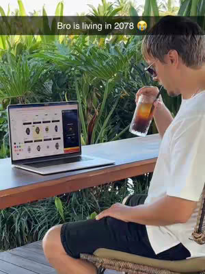 A man is seated outdoors, holding a drink and looking towards a laptop. He is wearing sunglasses and casual attire. The setting includes lush green foliage in the background.