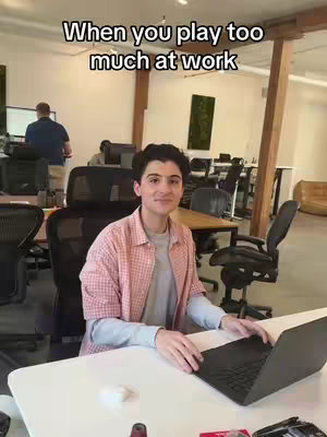 A man sits at a desk in an office setting, looking towards the camera with a slight smile. The background includes other office desks and individuals in the background.