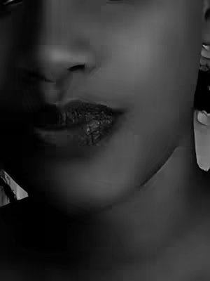 A close-up, black and white image presents a portion of a person's face. The focus is on the lower part of the face, highlighting the lips and the area around the nose. The lips appear dark and textured. The skin has a smooth appearance with shadows and highlights that create depth. The lighting seems soft and diffused, and the composition is simple and intimate.