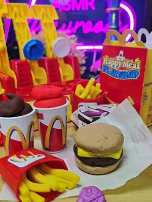 The image showcases a Play-Doh recreation of a McDonald's Happy Meal setting. It features Play-Doh burgers, fries, soft drink cups, and the iconic Happy Meal box. The background includes a Play-Doh version of a McDonald's seating area. The scene is brightly lit with a purple neon sign visible.