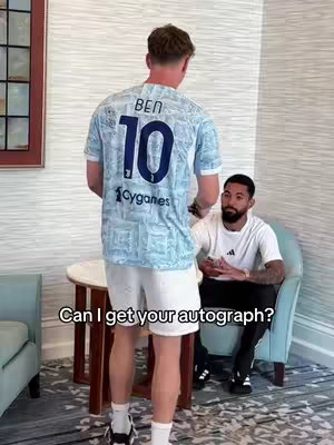 An interior shot shows two men interacting. The man in the foreground is wearing a white shirt and shorts, and a light blue jersey. From the perspective, the back of the jersey is facing the camera. The jersey has the name 'BEN' printed on the upper back, along with the number '10'. In front of him, is a man sitting down. Below the images are the words 'Can I get your autograph?'