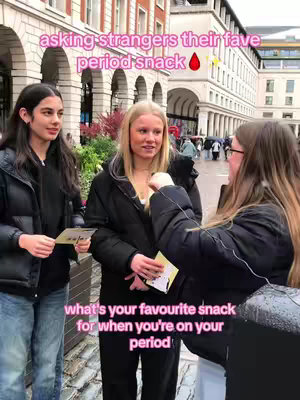 Three young women are standing on a city street talking. The woman on the right is holding a microphone and speaking to the other two, who are holding small cards. Text overlays on the image read "asking strangers their fave period snack" and "what's your favourite snack for when you're on your period". The background shows various buildings and a slightly overcast sky, which suggests a daytime setting.