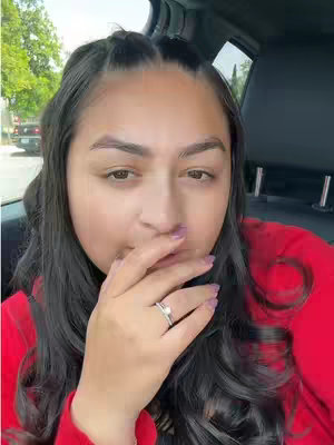A young woman is seated inside a car, with a hand covering her mouth and a thoughtful expression. The setting appears to be a sunny day, given the natural lighting. The composition is a close-up, focusing on her face and hand. The color palette is dominated by natural skin tones, the red of her shirt, black car interiors, and the clear sky outside.